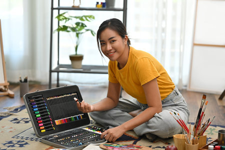 Portrait of A young Asian female artist sits on the floor, selecting colors from her art kit,...