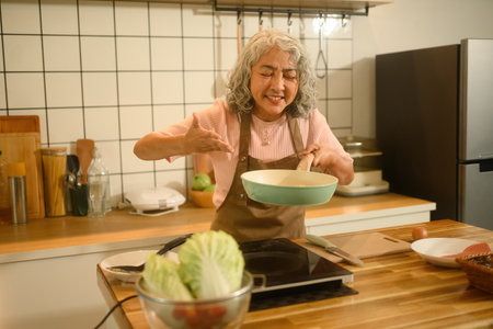 Smiling senior Asian woman in an apron cooking at home, holding a frying pan and presenting her food with joy in a cozy kitchen.の写真素材