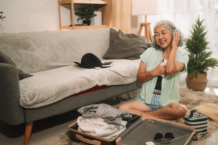 An elderly woman, sitting beside an open suitcase, enjoys music on headphones while packing for a trip in her cozy living room.の写真素材