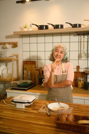 An elderly woman in an apron, enjoying music with headphones while cooking, holding a wooden spoon and smiling happily in her kitchen.の写真素材