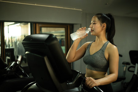 An Asian Woman Drinking Water on Treadmill at Gym, Hydration During Cardio Exercise.