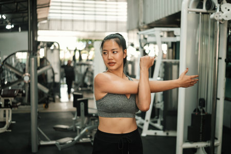 Confident young woman stretching her arm muscles in a gym, focusing on flexibility, strength, and...