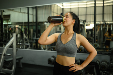 Confident young woman in sportswear enjoying a protein shake after a workout in a gym. Promotes fitness, healthy lifestyle, and post-exercise recovery.の写真素材