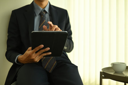 Close-up of a businessman in formal attire using a digital tablet, seated by the window with soft daylight.の写真素材