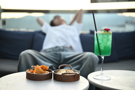 Refreshing green cocktail with snacks served on a rooftop table, with a relaxed man lounging in the...