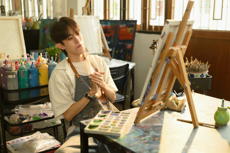 A young artist in a apron sits in a sunlit studio, thoughtfully looking at a canvas while surrounded...