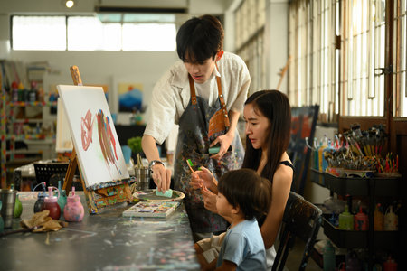 An art teacher guides a mother and child during a creative painting session in a colorful studio. Hands-on family learning and bonding through expressive, fun art activities.の写真素材