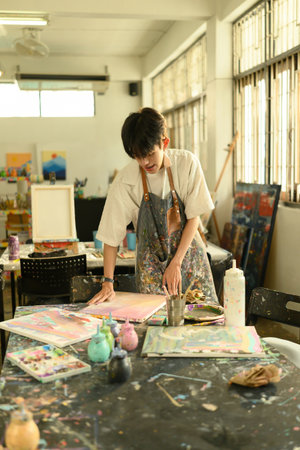 A young man in an apron engages with his colorful artwork in a bright studio setting. Art supplies, canvases, and creative tools fill the vibrant workspace.の写真素材