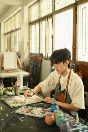 Vertical image of a Young male artist concentrating on painting with a brush at a table filled with colorful paints and tools in a bright, creative studio environment..の写真素材
