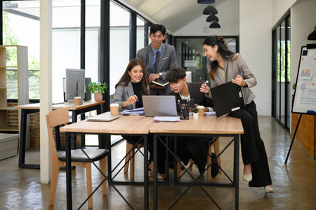 Group of young business professionals engaged in a lively brainstorming session around a laptop in a...