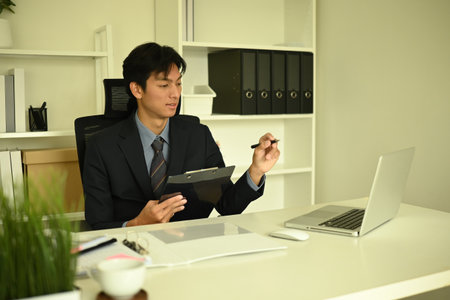 Young businessman in a suit sitting at a modern desk, holding a clipboard and pen, presenting or...