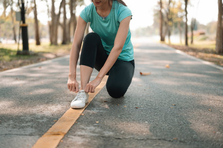 Close-up of a young Asian woman kneeling on a park path, tying her sneakers before starting a...