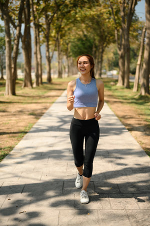 Happy young woman jogging on a shaded pathway in a park, enjoying an outdoor fitness routine in...