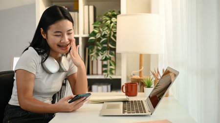 A cheerful Asian teenage girl chats with friends using her smartphone while sitting at a desk with a...