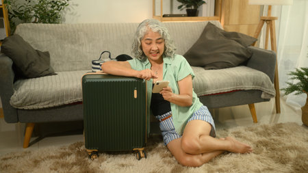 An Elderly Asian woman sitting next to her suitcase using a smartphone to book a hotel or confirm, and check tourist attraction reviews before traveling.の写真素材