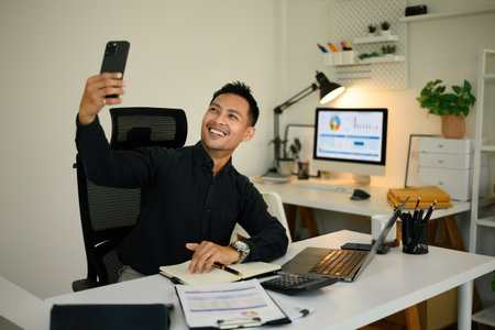 A cheerful businessman sitting at his desk, smiling and holding a smartphone during a video call in...