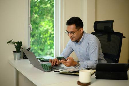 A mature Asian businessman in a clean modern office uses his smartphone while sitting at a desk with...