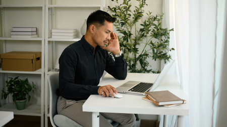 A thoughtful businessman sits at a desk, using a computer with a focused expression.