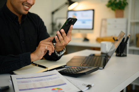 Close-up of a businessman using a smartphone at his desk, with financial documents, a laptop, and a...