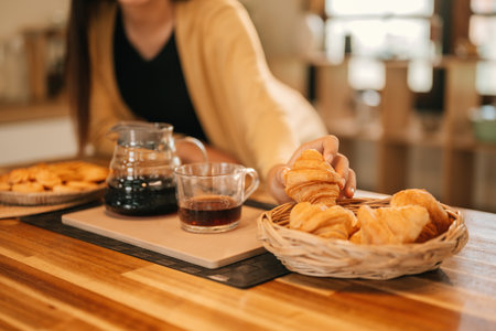 Close-Up of A woman's hand reaches for a golden croissant from a basket, paired with a glass of black coffee on a wooden kitchen counter.の写真素材