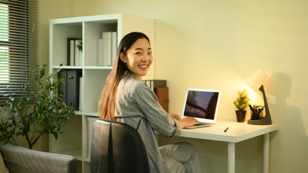 Confident young woman smiling at the camera while working at her home office desk with a laptop.