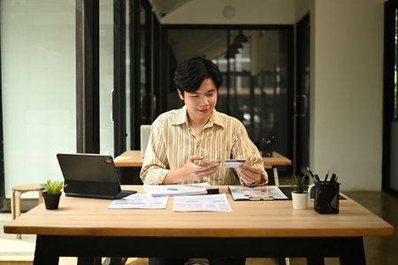 Young Asian man makes an online payment using a smartphone and credit card at a modern office desk.
