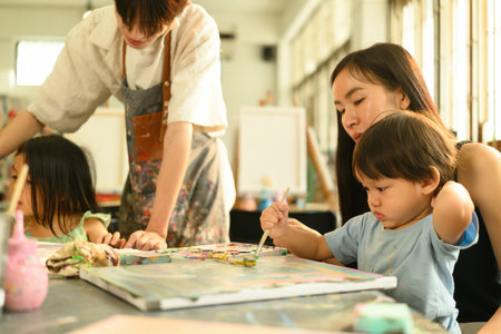 A young boy concentrates on his painting, guided by his mother and watched over by a supportive art...