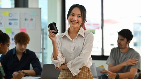 Confident young Asian woman holding a coffee cup while smiling in a office workspace with team...