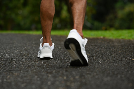 Close-up of athletic manâ€™s legs and white running shoes walking on an outdoor park pathway.