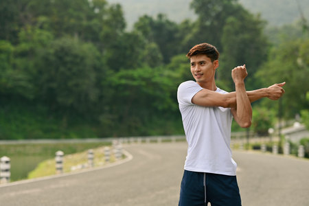 A Fit young Asian man preparing for a run, stretching muscles outdoors on a scenic road.