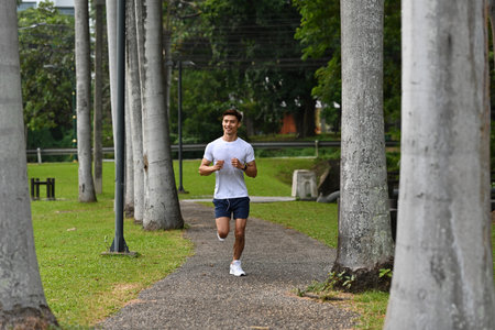 An Asian man jogging on a scenic park trail lined with tall trees, enjoying outdoor exercise and...