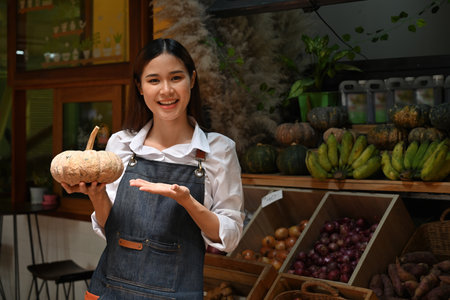 Young female vendor presenting a fresh pumpkin at a vegetable market, surrounded by local produce.の写真素材