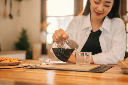 Close-up of a woman pouring a black coffee from a glass pot into a clear cup on a wooden kitchen counter.の写真素材