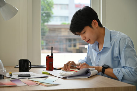 Young architect working intently at a modern desk, using a ruler and pen to measure blueprints...