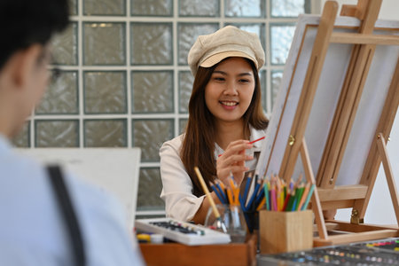 An Asian woman artist in a beige hat and apron smiles as she paints on canvas beside another painter...