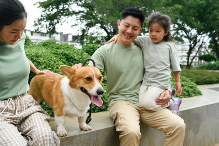 A father holds his daughter while petting his Corgi with his wife sitting beside him amidst a green...