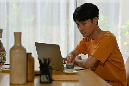 A handsome Asian man in casual outfit focused on working on a laptop in a modern living room. for...