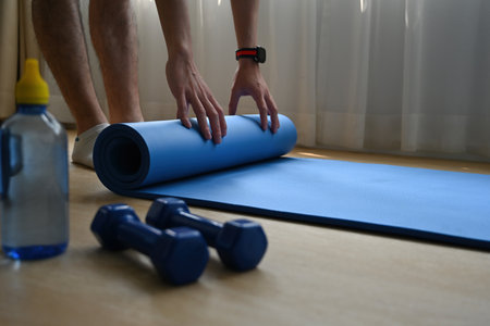 A cropped view of bottom part of a man rolling the yoga mat in a studio with the dumb bells and...