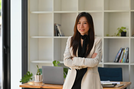 A portrait of a young pretty Asian woman standing in the office, crossing arms smiling, for real estate, business, finance and technology concept.の写真素材