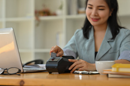 Cropped view of a smiling Asian woman sitting in the office holding a credit card and using a credit...