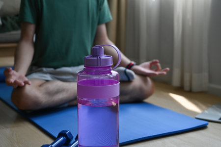 Close up focused water bottle in the foreground and a man sitting cross-legged meditating on a mat...
