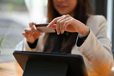 Cropped view of a smiling Asian woman wearing a suit sitting in the office holding a credit card...