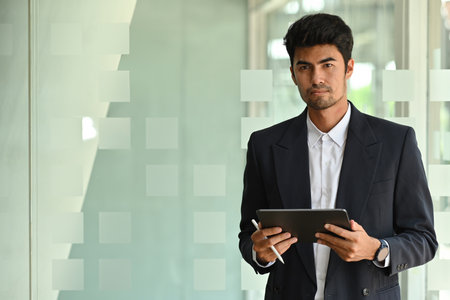 A portrait of a handsome Asian businessman standing in a workplace holding a tablet and looking at the camera, for business and technology concept.の写真素材