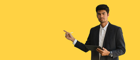 The confident Indian Asian businessman in suit pointing a finger for presentation and holding a digital tablet over a yellow background. Presentation for business. Business and financial concepts.の写真素材