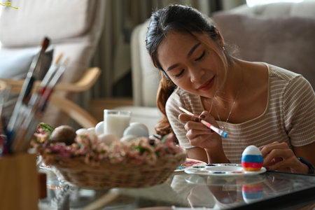 Asian beautiful female looking thoughtful while painting an Easter egg on color palette with Easter holiday decoration on table.の写真素材