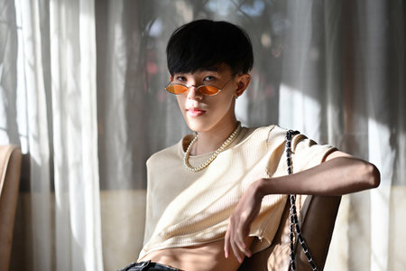 Portrait of LGBT Asian slim male with crop top cream shirt and orange glasses looking at camera and sitting on chair with a pose and sunlight. People lifestyle fashion LGBTQ concept.の写真素材