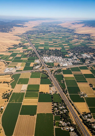 Aerial photograph of green and brown patchwork fields, highways, and buildings under clear blue sky