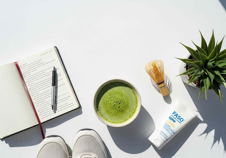 Flat lay of green tea, notebook, pen, shoes, plant, and skincare products on white surface