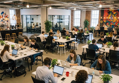 Collaborative workspace with numerous people working on laptops at shared white tables with plants