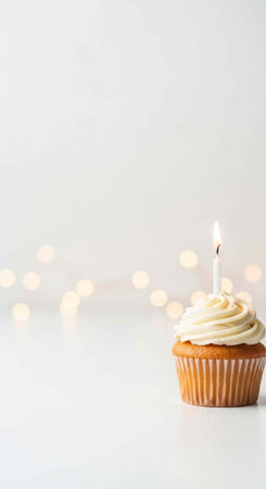 A single cupcake with white frosting and a lit candle on top, against a white background with blurred lightsの写真素材